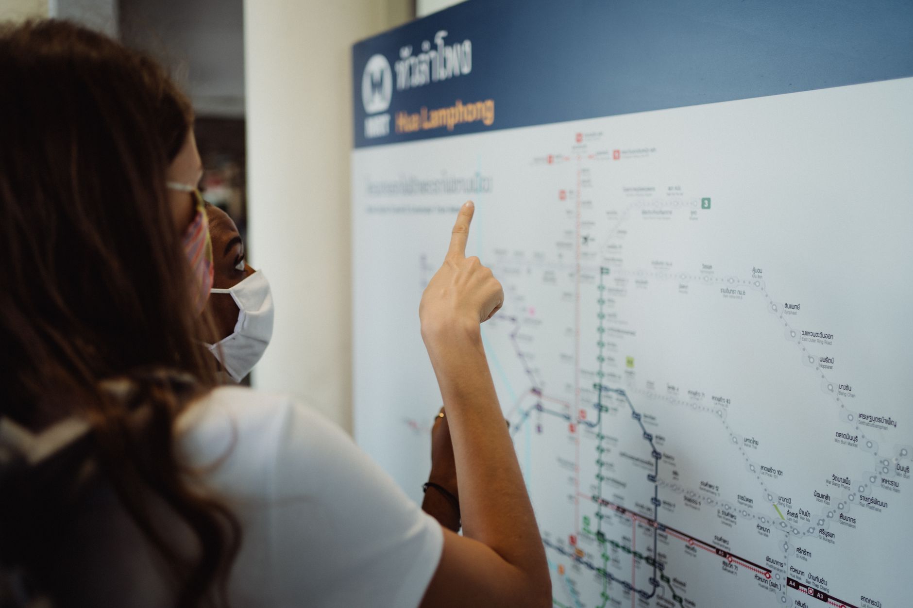 Two women in face masks pointing at a transport map, exploring new directions. — Backpackers Travel Cover - An Inexpensive Al