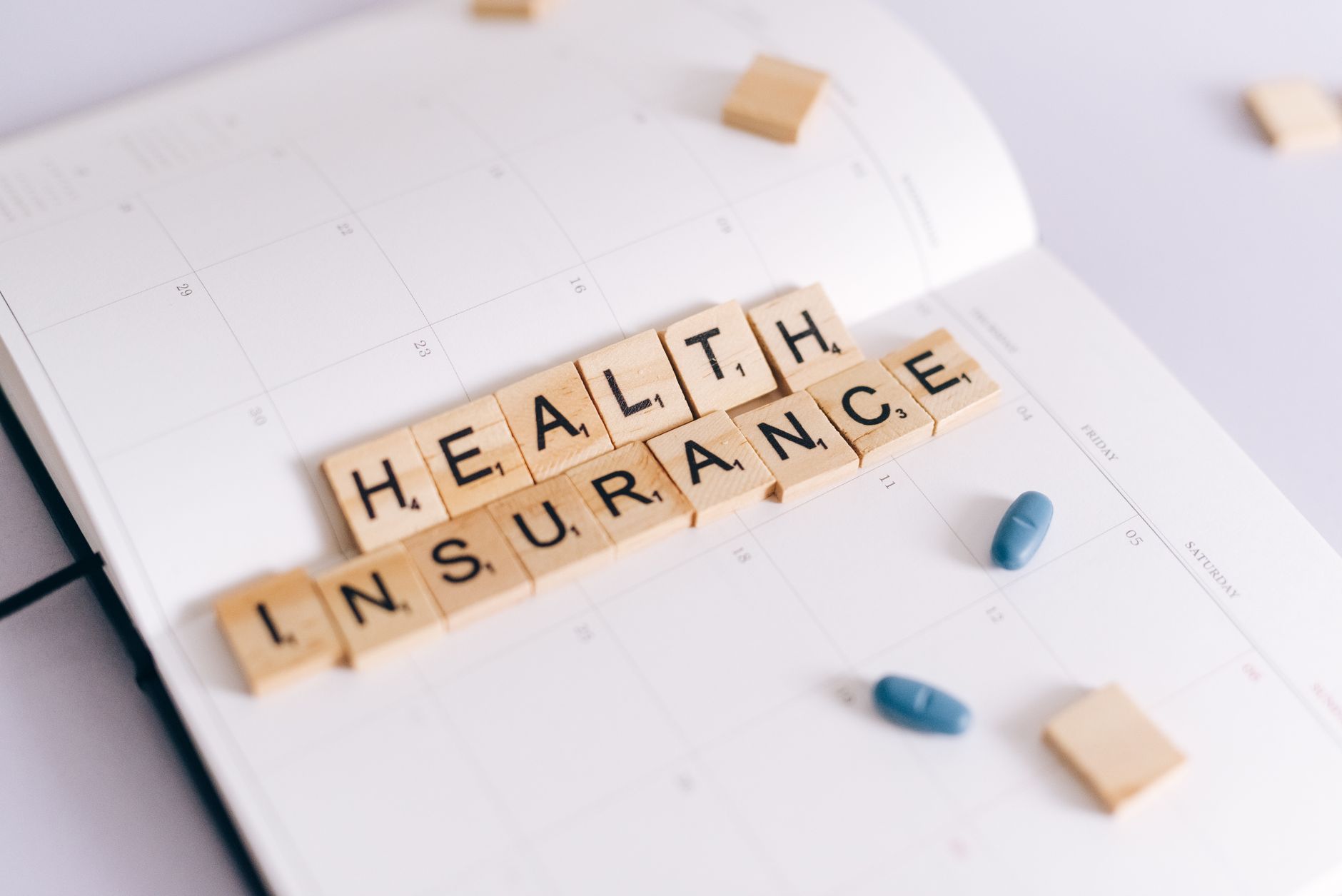 Close-up of health insurance spelled out with Scrabble tiles on a planner, alongside medication pills. — Yo-Yo Health Habits